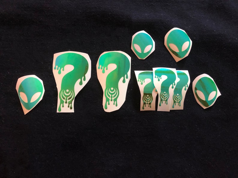 May include: A set of nine green and white iron-on patches featuring alien heads and question marks. The patches are perfect for adding a unique touch to clothing, bags, or other accessories.
