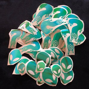 May include: A pile of green and blue holographic stickers featuring alien head and dripping liquid designs.
