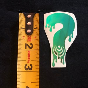 May include: A green and blue iridescent vinyl sticker of a dripping question mark with a stylized design inside. The sticker is about 2 inches tall.