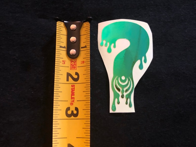 May include: A green and blue iridescent vinyl sticker of a dripping question mark with a stylized design inside. The sticker is about 2 inches tall.
