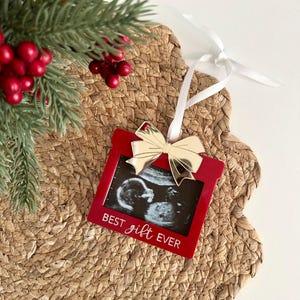 Op de afbeelding: Rode kerstversiering met een echografiebeeld, een gouden strik en de tekst "BEST gift EVER". De versiering hangt aan een wit lint. Het rust op een geweven mat, met een dennentak en rode bessen op de achtergrond.