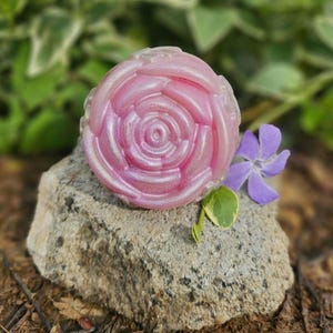 May include: A pink rose-shaped soap bar with a shimmery finish, resting on a gray rock with a purple flower.
