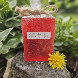 May include: A rectangular red glycerin soap bar, wrapped in clear cellophane and tied with twine. The label reads "Scarlet Petals Glycerin Soap." A yellow dandelion flower rests on a rock next to the soap.