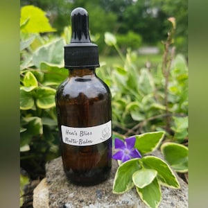 Könnte beinhalten: Eine braune Glasflasche mit schwarzem Tropfaufsatz, beschriftet mit "Hen's Bliss Multi-Balm". Die Flasche steht auf einem Stein, umgeben von grünem und weißem Laub und einer kleinen lila Blume. Der Hintergrund ist unscharf.