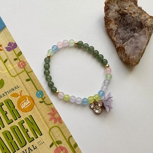 May include: A multi-colored beaded bracelet with a flower charm and a gold accent. The beads are green, blue, pink, and yellow.