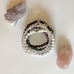 May include: A set of four stacked bracelets made of amethyst, green aventurine, and white howlite beads. The bracelets are accented with silver beads and a silver charm.