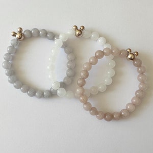 May include: Three beaded bracelets in shades of gray, white, and tan. Each bracelet features a spherical bead design and a silver-colored Mickey Mouse head charm. The bracelets are displayed on a white surface.