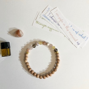 May include: A wooden beaded bracelet with gold accents and various colored stones. The bracelet is on a white surface with several cards with handwritten affirmations.