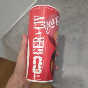 May include: A red and white cylindrical plastic cup featuring the text Collector's Edition, GER v CIV, Coca-Cola, and FIFA Official Partner. The design includes graphic elements in red, white, and yellow, with a smooth, glossy finish throughout.