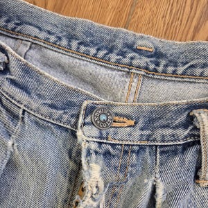May include: Close-up of light blue denim jeans with a button that reads "EDWIN CO. LTD". The jeans have distressed details, including frayed edges and a worn appearance. The stitching is visible in a contrasting tan color.