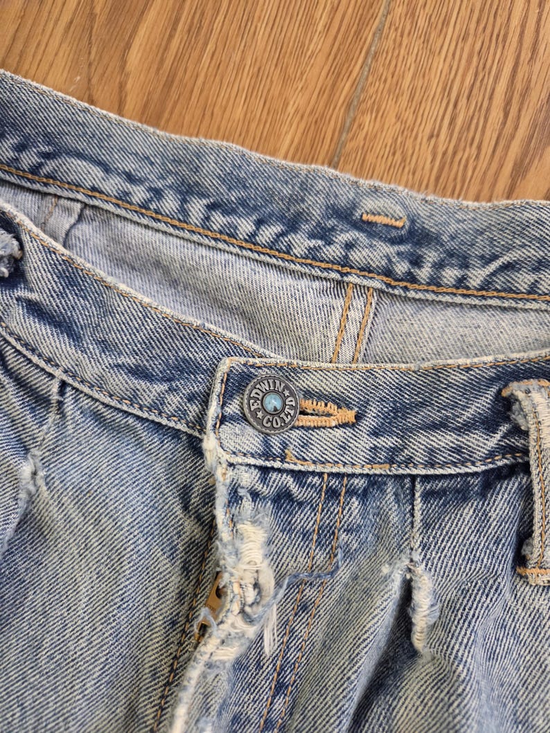 May include: Close-up of light blue denim jeans with a button that reads "EDWIN CO. LTD". The jeans have distressed details, including frayed edges and a worn appearance. The stitching is visible in a contrasting tan color.