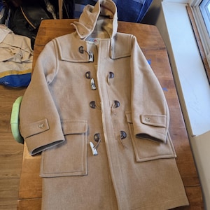 May include: A tan duffel coat with a hood, toggle closures, and two front pockets. The coat is made of a thick, wool-like material and is laid out on a wooden surface. The coat has a classic, timeless design.