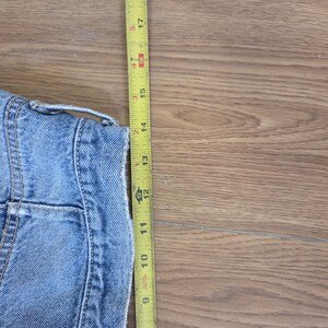 May include: A light blue denim jacket with a yellow measuring tape. The jacket's length is approximately 14 inches. The tape shows measurements in inches, with the jacket's details visible.