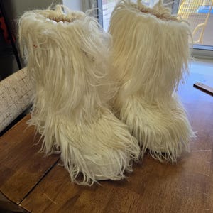 Puede incluir: Un par de botas peludas de color blanquecino. Las botas est&aacute;n hechas de un material largo y esponjoso, lo que les da un aspecto texturizado. Est&aacute;n sobre una superficie de madera.