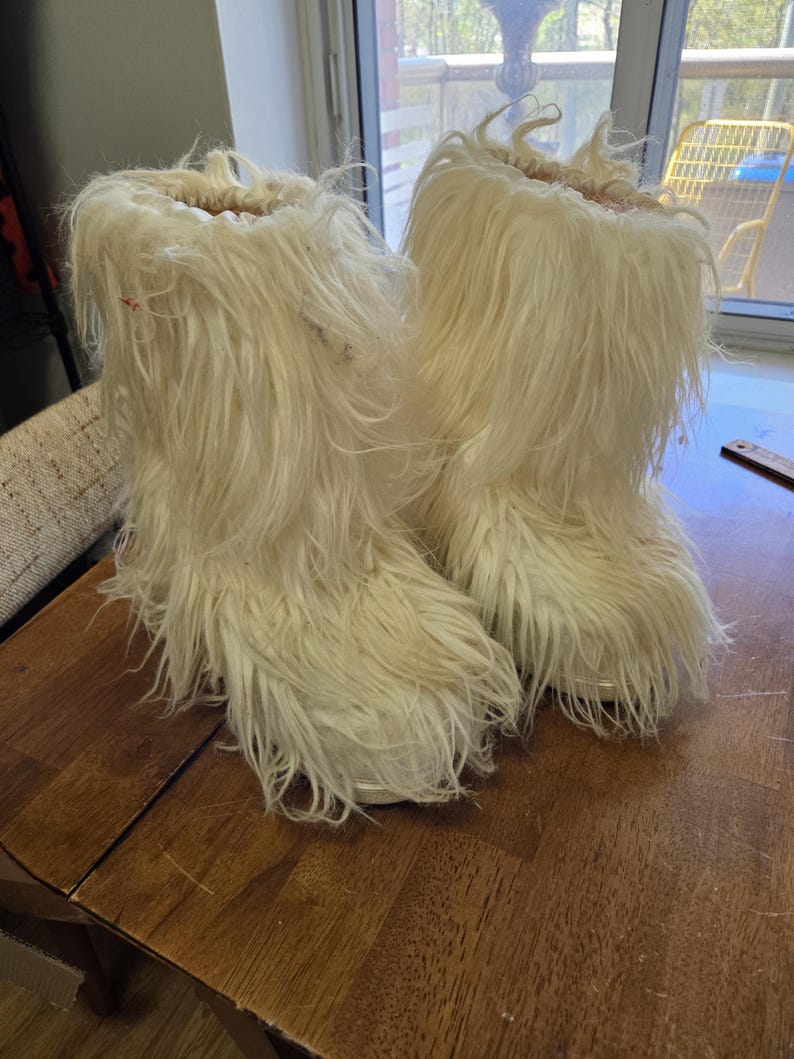 Puede incluir: Un par de botas peludas de color blanquecino. Las botas est&aacute;n hechas de un material largo y esponjoso, lo que les da un aspecto texturizado. Est&aacute;n sobre una superficie de madera.