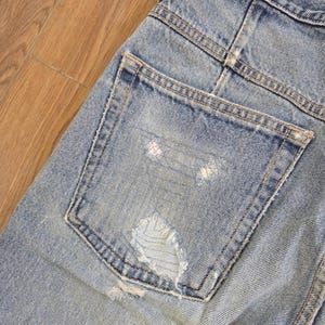 May include: Close-up of light blue denim jeans with a distressed pocket. The jeans show signs of wear with a ripped section on the back pocket. The stitching is visible, and the denim has a faded appearance.