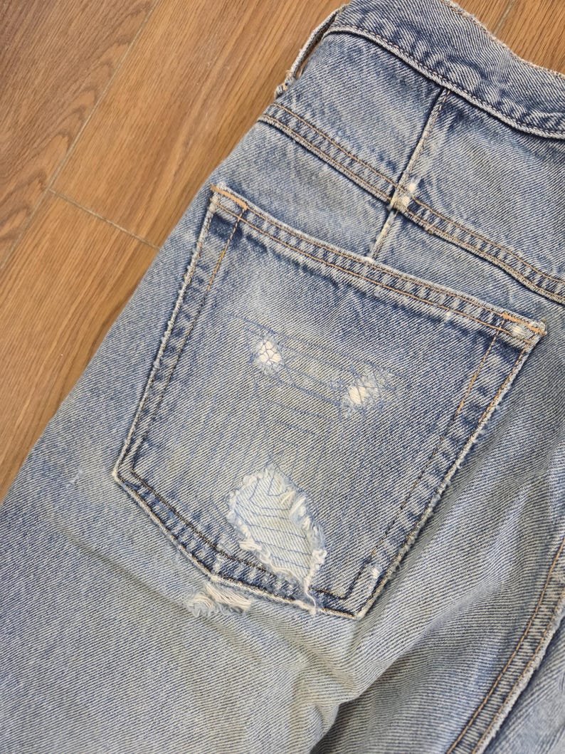 May include: Close-up of light blue denim jeans with a distressed pocket. The jeans show signs of wear with a ripped section on the back pocket. The stitching is visible, and the denim has a faded appearance.