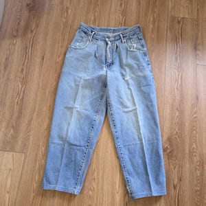 May include: Light blue denim jeans with a high waist and a relaxed fit. The jeans have a button and zipper closure, front pockets, and a few distressed areas. The pants are laid flat on a wooden surface.
