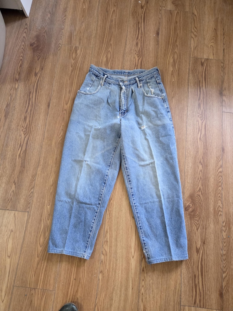 May include: Light blue denim jeans with a high waist and a relaxed fit. The jeans have a button and zipper closure, front pockets, and a few distressed areas. The pants are laid flat on a wooden surface.