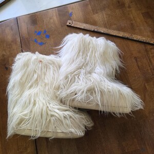 Puede incluir: Un par de botas peludas de color blanquecino. Las botas tienen aspecto de piel sint&eacute;tica, con fibras largas y fluidas. Las suelas son de un color m&aacute;s claro y las botas parecen ser de altura de tobillo. Las botas est&aacute;n sobre una superficie de madera.