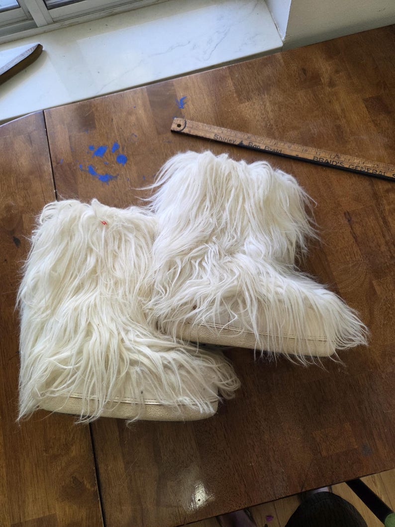 Puede incluir: Un par de botas peludas de color blanquecino. Las botas tienen aspecto de piel sint&eacute;tica, con fibras largas y fluidas. Las suelas son de un color m&aacute;s claro y las botas parecen ser de altura de tobillo. Las botas est&aacute;n sobre una superficie de madera.
