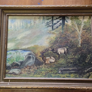 May include: An oil painting in a gold frame depicts a forest scene with a pond and two elk. The elk are brown and white, one standing and one lying down. The painting has a vintage aesthetic.