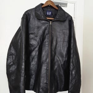 May include: Black leather jacket with a zip-up front, hanging on a wooden hanger. The jacket has a collar and the brand name "GAP" is visible on the label. The jacket is hanging on a white door.