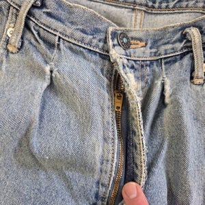 May include: Close-up of light blue denim jeans with a partially unzipped brass zipper. The jeans show signs of wear and tear, with visible fading and distressed details. The button is visible, and the jeans are on a wooden surface.