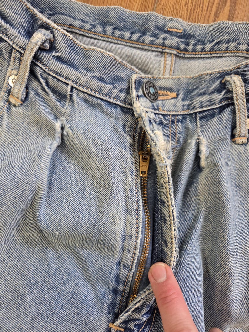 May include: Close-up of light blue denim jeans with a partially unzipped brass zipper. The jeans show signs of wear and tear, with visible fading and distressed details. The button is visible, and the jeans are on a wooden surface.