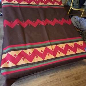 May include: A large brown blanket featuring horizontal bands with a repeating pattern of red zigzags and tan triangles. The design includes thin green and black stripes bordering the geometric sections, creating a symmetrical layout across the fabric.