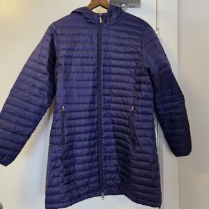 May include: A long, quilted, navy blue jacket with a hood and a full-length zipper. The jacket has two zippered pockets on the front and is hanging on a wooden hanger. The jacket is made of a lightweight material.