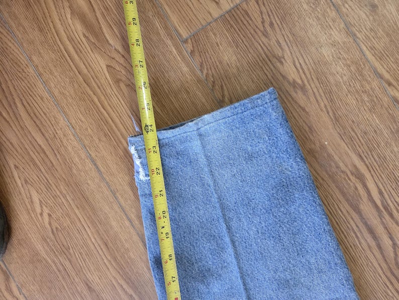 May include: A pair of light blue denim jeans with a yellow measuring tape showing a length of approximately 23 inches. The jeans are folded, and the tape is positioned along the edge, indicating a measurement.