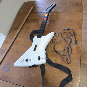 May include: A white video game guitar controller with a black neck, five coloured fret buttons, a black strum bar, and a black shoulder strap. The controller features a wired USB connection cable and a white body with a distinct angular shape.