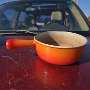Peut inclure: Une casserole en fonte émaillée orange et rouge avec une seule poignée. La casserole a un intérieur blanc et repose sur une surface rouge.