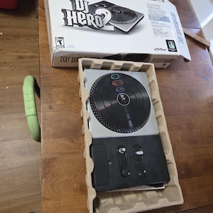 May include: A DJ Hero 2 video game turntable controller for the PS3 console, featuring a black circular platter with colored buttons and a black control panel with silver sliders, housed in a molded cardboard tray with its original white box.