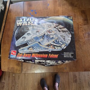 May include: A rectangular cardboard box for an AMT Star Wars Cut-Away Millennium Falcon model kit. The box features a graphic of the spacecraft with removable sections showing the inner structure, set against a dark space background with a planet.