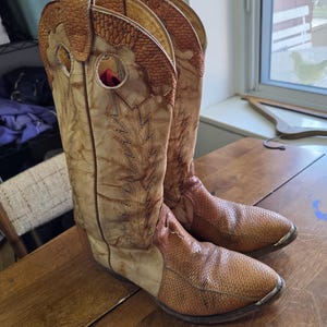 May include: A pair of vintage cowboy boots. The boots are tan and brown with decorative stitching and a textured pattern on the toe and upper shaft. The boots are made of leather and have a classic Western design.