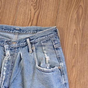 May include: Close-up of light blue denim jeans with a faded wash and distressed details. The jeans feature a button closure, belt loops, and a classic five-pocket design. The fabric shows signs of wear and tear, adding to the vintage aesthetic.