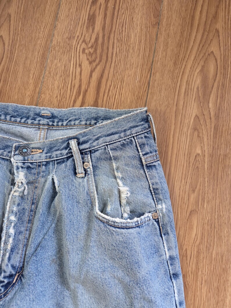 May include: Close-up of light blue denim jeans with a faded wash and distressed details. The jeans feature a button closure, belt loops, and a classic five-pocket design. The fabric shows signs of wear and tear, adding to the vintage aesthetic.