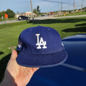 May include: A royal blue baseball cap featuring the white embroidered LA logo on the front crown. The cap has a flat brim and includes a small embroidered patch on the side panel. The design is structured with a button on top and eyelets for ventilation.