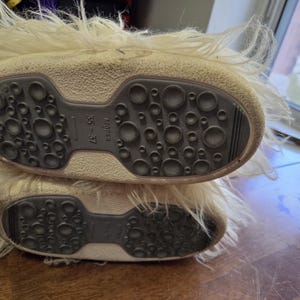 Puede incluir: Un par de botas de invierno color crema con suela texturizada gris oscuro con agarres circulares. Las botas tienen un ribete de material esponjoso, similar a la piel. La talla indicada es 35-37. La suela mide unos 23 cm.