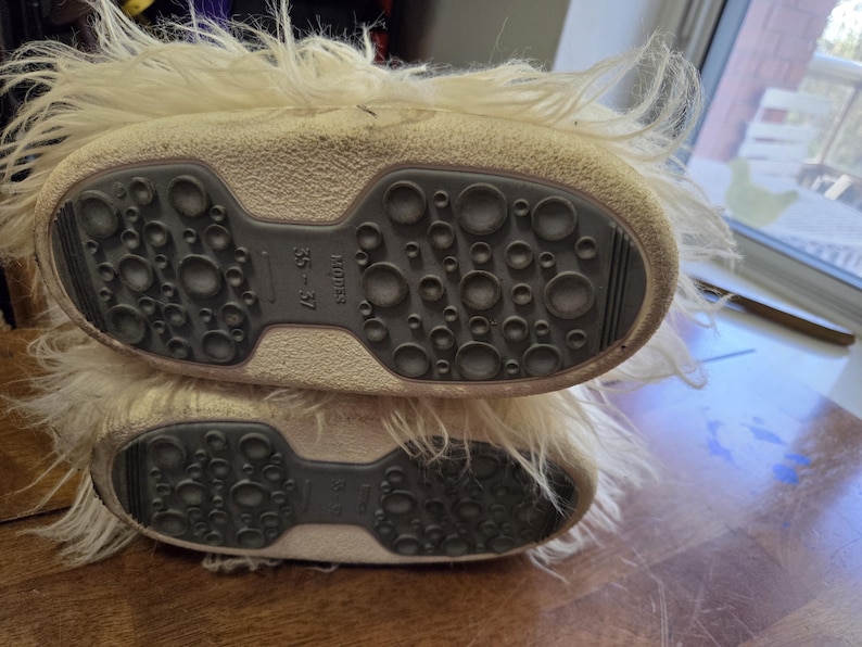 Puede incluir: Un par de botas de invierno color crema con suela texturizada gris oscuro con agarres circulares. Las botas tienen un ribete de material esponjoso, similar a la piel. La talla indicada es 35-37. La suela mide unos 23 cm.