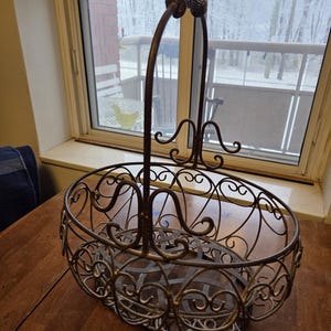 May include: An oval metal basket in an antique style with a handle. The basket features an ornate, openwork design with swirling patterns and a bronze finish. The arched handle is topped with decorative spheres. The basket is on a wooden table near a window.
