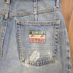 May include: Close-up of light blue denim jeans with a distressed pocket. The pocket features a rectangular patch with the brand name "EDWIN" and other text. The jeans show signs of wear and tear.