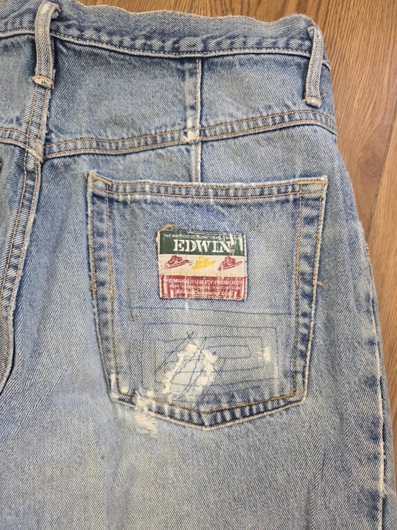 May include: Close-up of light blue denim jeans with a distressed pocket. The pocket features a rectangular patch with the brand name "EDWIN" and other text. The jeans show signs of wear and tear.