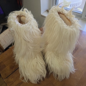 Puede incluir: Un par de botas peludas de color blanquecino. Las botas tienen una textura suave y peluda y un interior de color claro. Las botas est&aacute;n dise&ntilde;adas para cubrir los pies y los tobillos, con un exterior esponjoso.