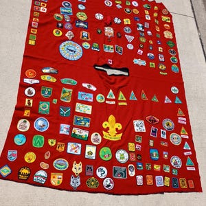Peut inclure: Une couverture en tissu rouge ornée de nombreux écussons colorés. Les écussons présentent divers motifs, notamment des badges, des logos et du texte. La couverture est posée sur une surface en béton.