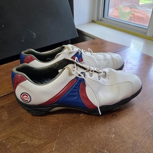 May include: A pair of white athletic shoes featuring blue and red side panels, white laces, and a circular Chicago Cubs logo on the heel. The shoes have a black sole and a low-top silhouette with a smooth exterior finish and metallic eyelets.