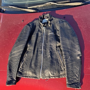 May include: Black leather jacket with a zipper closure, two zippered pockets, and a small pocket on the chest. The jacket has a stand-up collar and a worn, textured appearance. The jacket is displayed on a red surface.