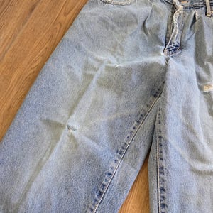May include: Light blue denim jeans with a faded wash and distressed details. The jeans have a classic five-pocket design, a zipper fly, and a button closure. The fabric shows signs of wear and tear, adding to the vintage aesthetic.